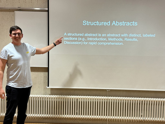 Prof. Dr. Sven Mayer stands next to a projection screen in a lecture room, smiling and pointing at a slide titled “Structured Abstracts.” The slide explains that a structured abstract contains labeled sections such as Introduction, Methods, Results, and Discussion.