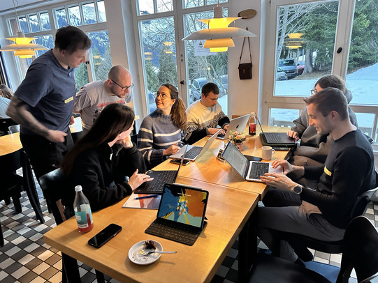 Several participants sit around a long wooden table in a bright room with large windows, working together on laptops and tablets. Some people are typing while others lean in to discuss their work. Drinks, notebooks, and a small plate with cake are on the table. Outside the windows, snow-covered trees and mountains are visible, creating a scenic backdrop for the collaborative workshop setting.