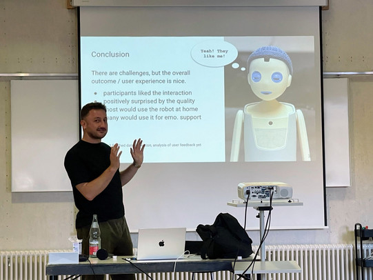 Matthias Schmidmaier stands in front of a projection screen in a lecture room, gesturing with both hands while presenting. The slide shows a conclusion section with a humanoid robot illustration and a speech bubble reading “Yeah! They like me!” The presentation is part of a talk on “Realtime Interaction with Intelligent Agents in User Research.” A table with a laptop, microphone, and projector is visible in the foreground, indicating an academic workshop setting.