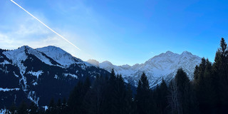 A wide panoramic view of snow-covered mountain peaks under a clear blue sky. Dark evergreen trees fill the foreground, while sunlight illuminates the distant ridges. A white airplane contrail stretches diagonally across the sky, adding a sense of motion to the calm alpine landscape.
