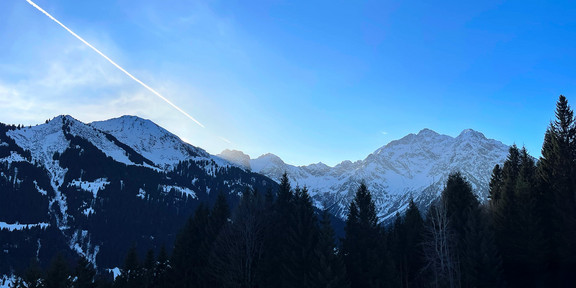 A wide panoramic view of snow-covered mountain peaks under a clear blue sky. Dark evergreen trees fill the foreground, while sunlight illuminates the distant ridges. A white airplane contrail stretches diagonally across the sky, adding a sense of motion to the calm alpine landscape.