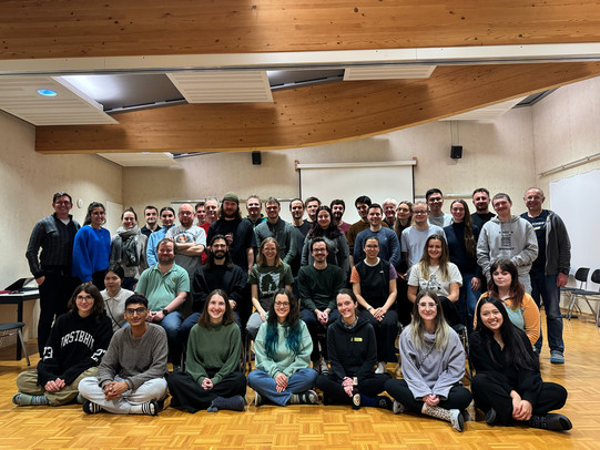 A large group of around fivty people pose for a group photo in a bright seminar room with wooden ceilings and parquet flooring. They are arranged in several rows, with some sitting on the floor in front and others standing behind. Most are smiling and looking at the camera. A projector screen and chairs are visible in the background, indicating an academic or workshop setting.