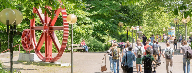 Studenten im Sommer auf dem Nordcampus, rote Zahnräder im Hintergrund.