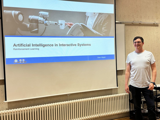 Prof. Dr. Sven Mayer stands smiling next to a large projection screen in a lecture room. The screen displays a presentation slide titled “Artificial Intelligence in Interactive Systems – Reinforcement Learning,” featuring an image of a robotic arm holding a cup. A radiator and chairs are visible below and beside the screen, indicating an academic teaching setting.